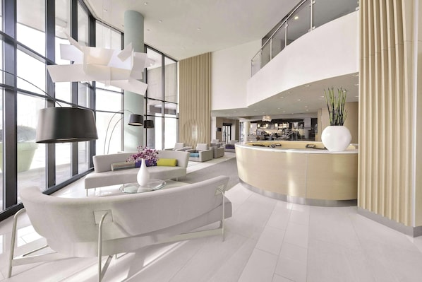 A modern hotel lobby with a curved reception desk, a seating area with sofas, and large windows offering views of the city.