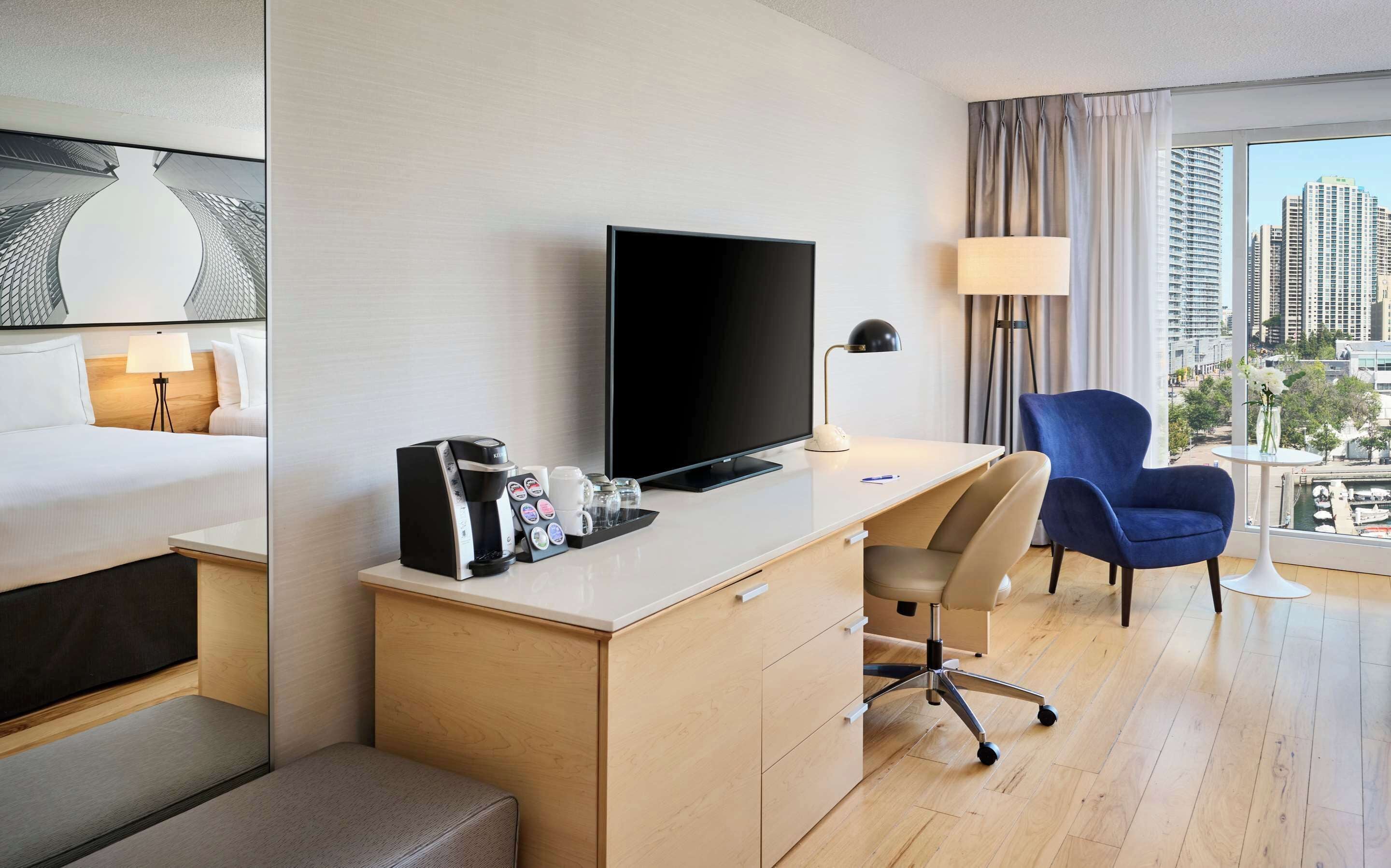 Hotelkamer met een bed, een bureau met koffiezetapparaat, een televisie, een stoel en uitzicht op de stad.