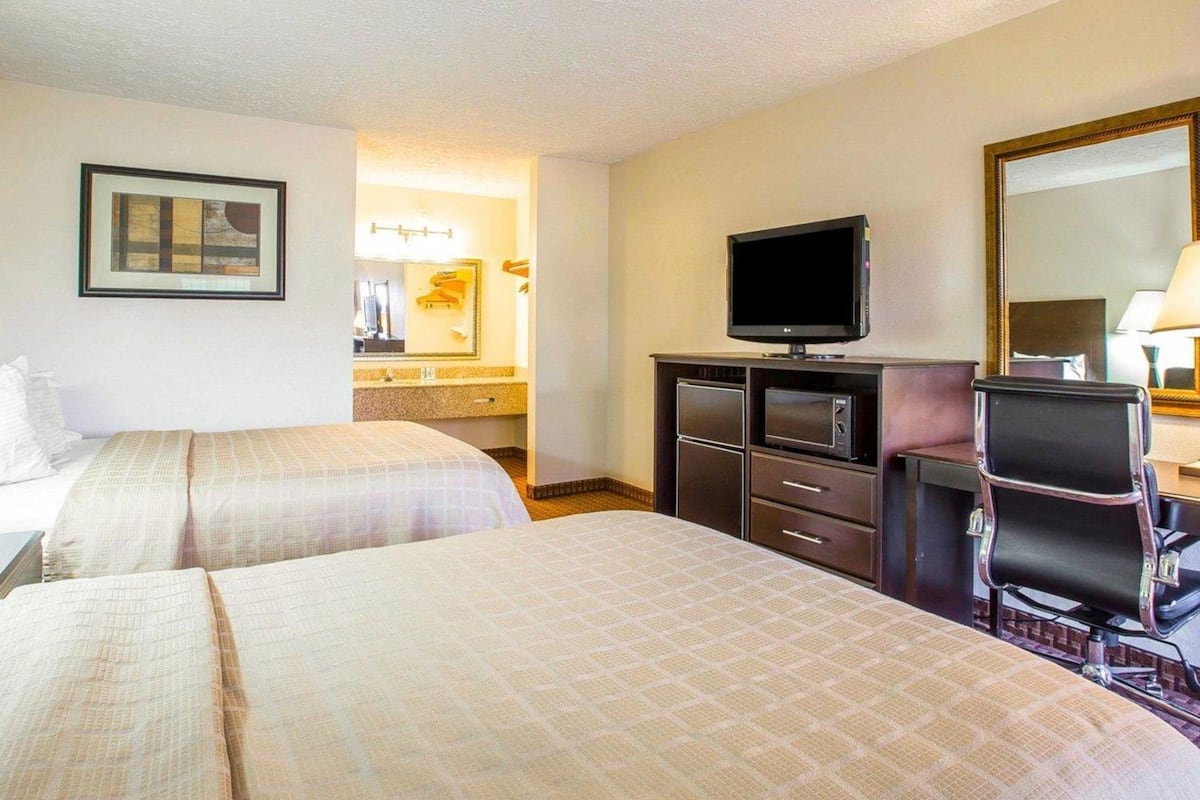 A hotel room with two beds, a television, a desk with a chair, a mirror, and a framed picture on the wall.