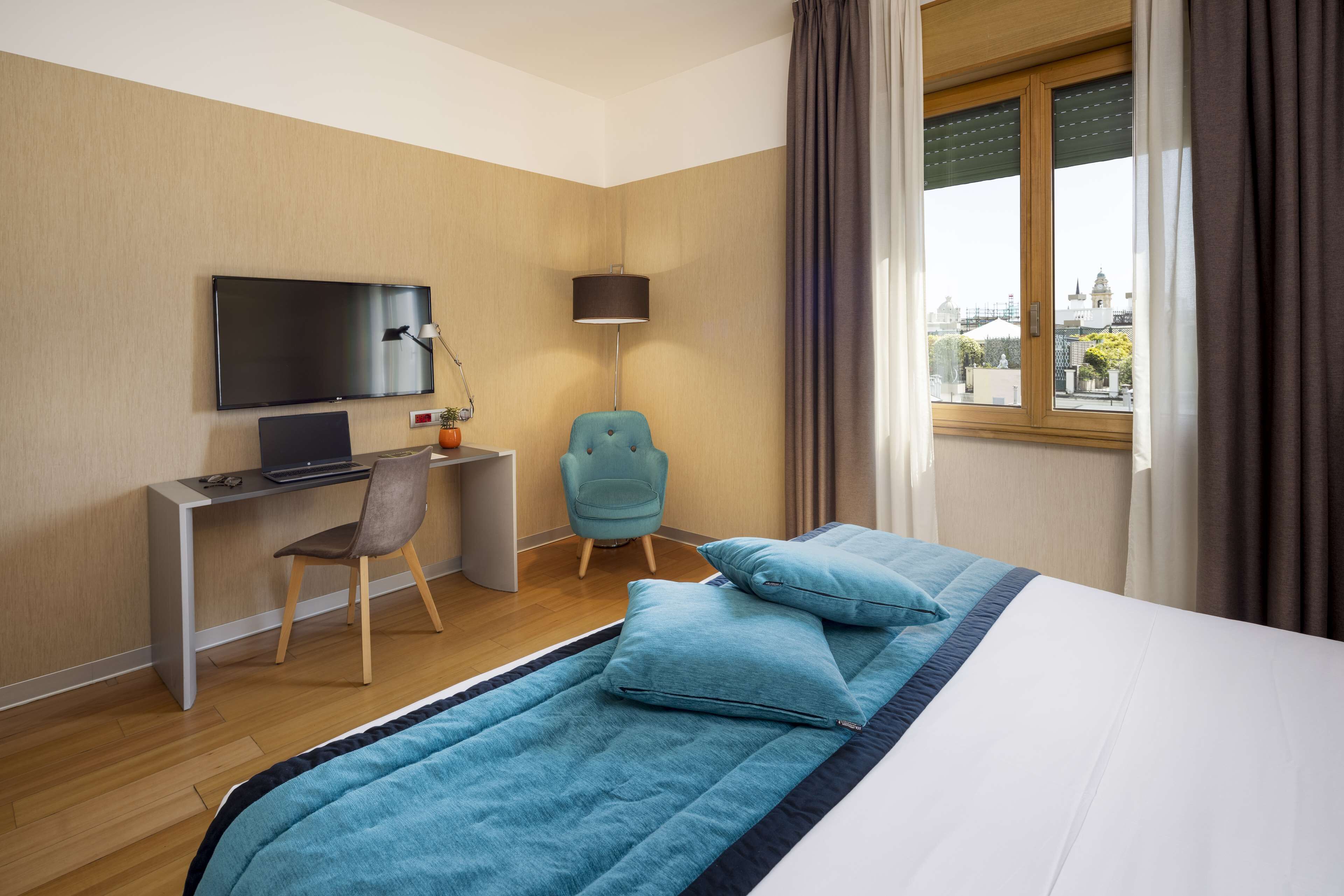 A hotel room with a bed, a desk with a computer, a chair, a TV, and a window with curtains.