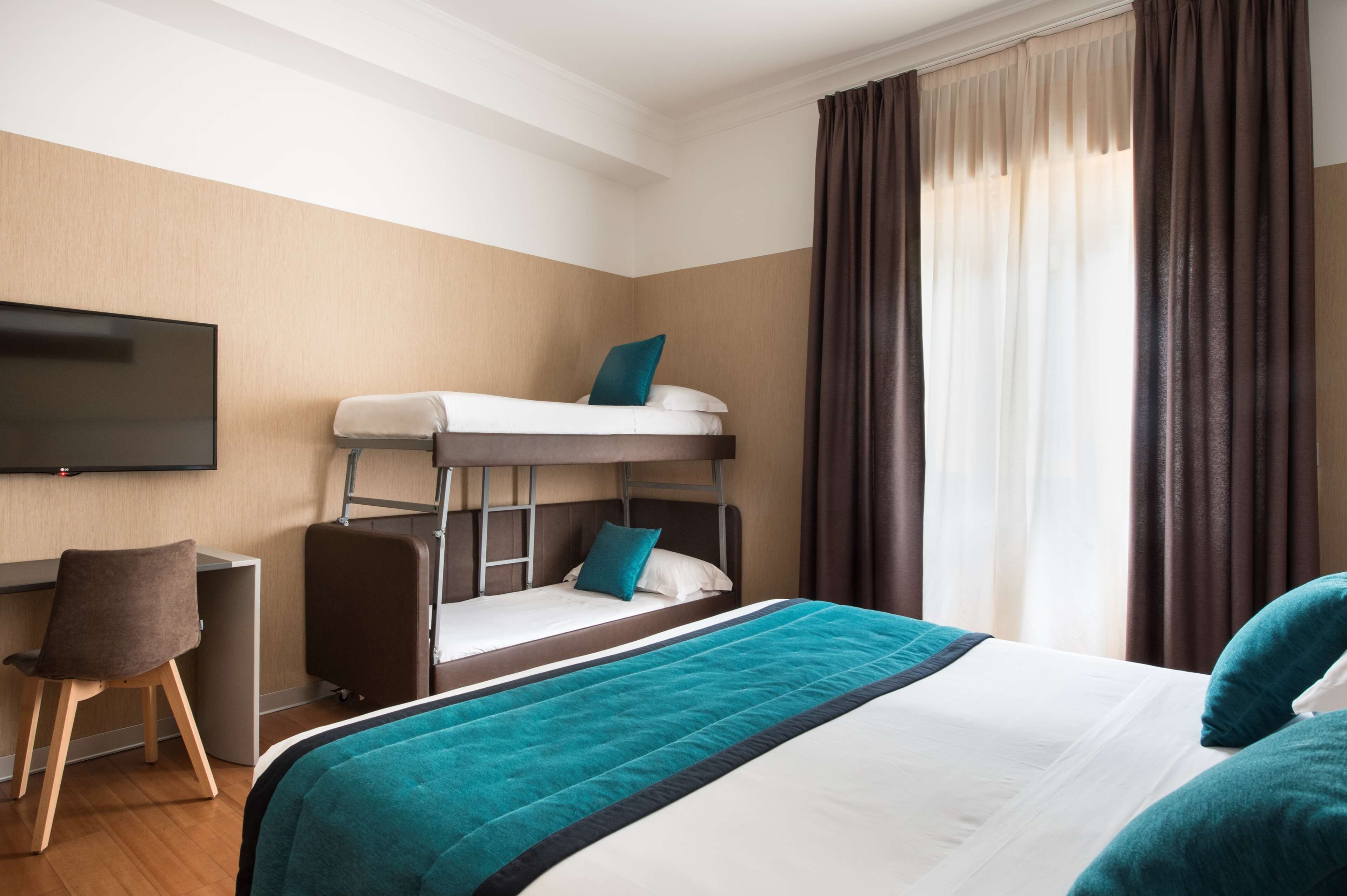 A hotel room with a bunk bed, a desk, a chair, a flat-screen TV, and a large window with curtains.