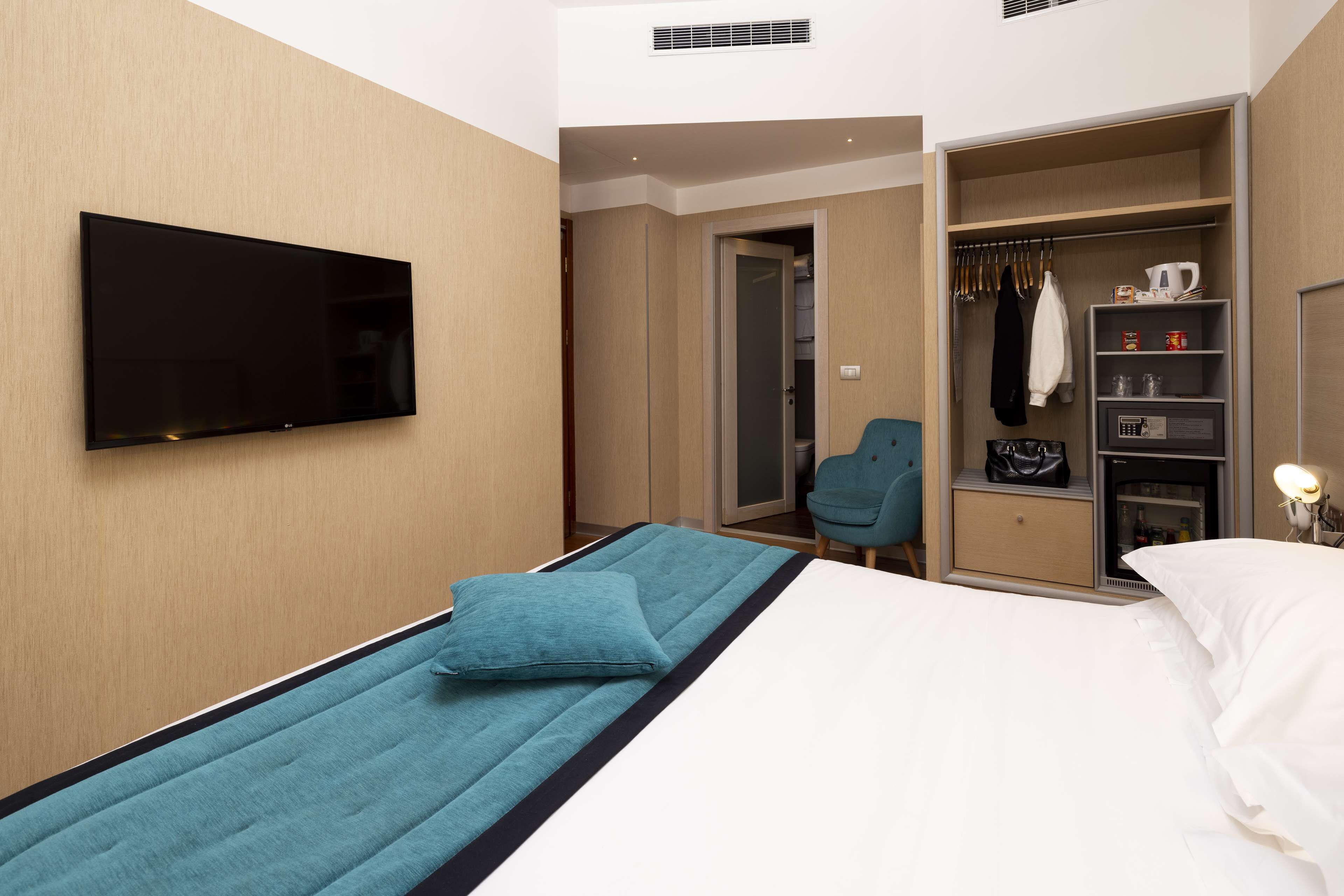 A hotel room with a bed, a TV mounted on the wall, a seating area, and a wardrobe with a mirror.
