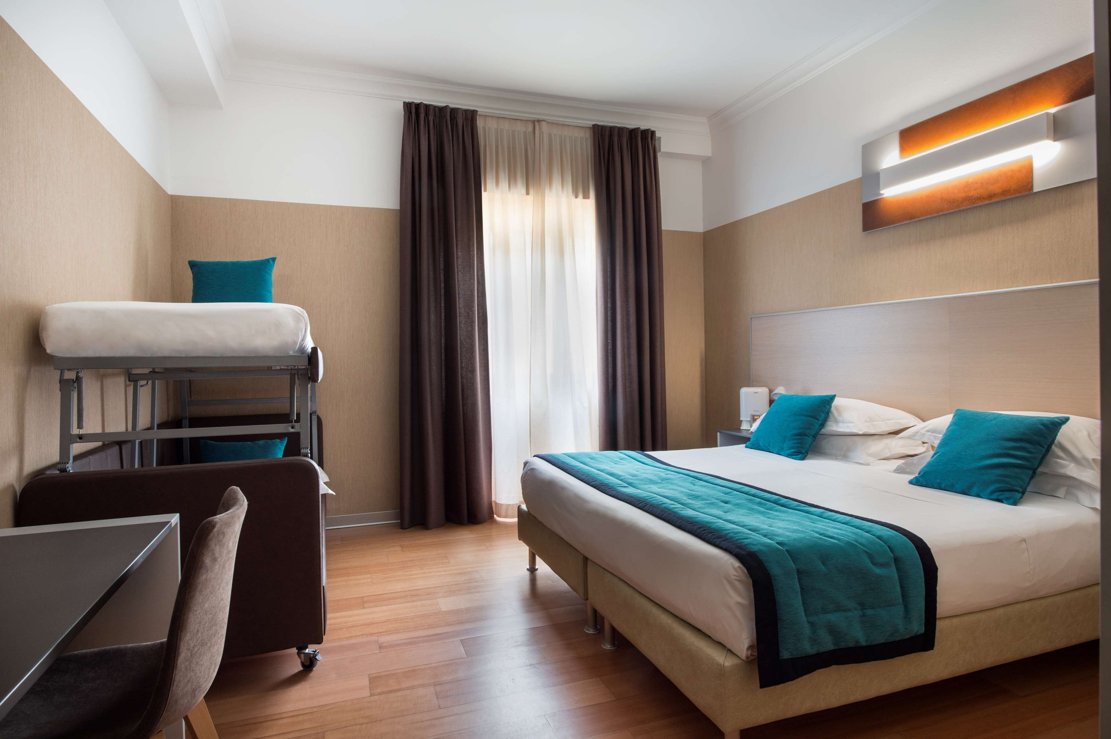 Une chambre d’hôtel avec un lit superposé, un bureau avec une chaise, un lit recouvert d’une couverture turquoise et une table de chevet.