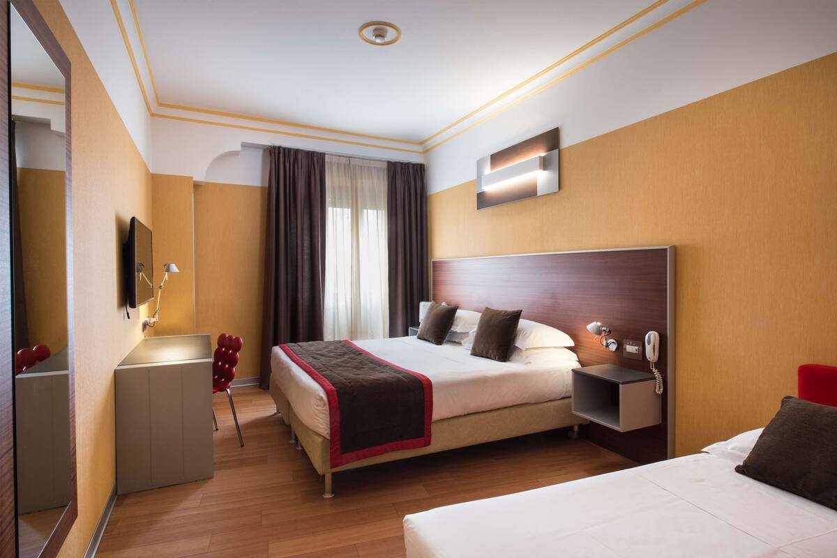 A hotel room with a large bed, bedside tables, a mirror, a desk, and a red chair.