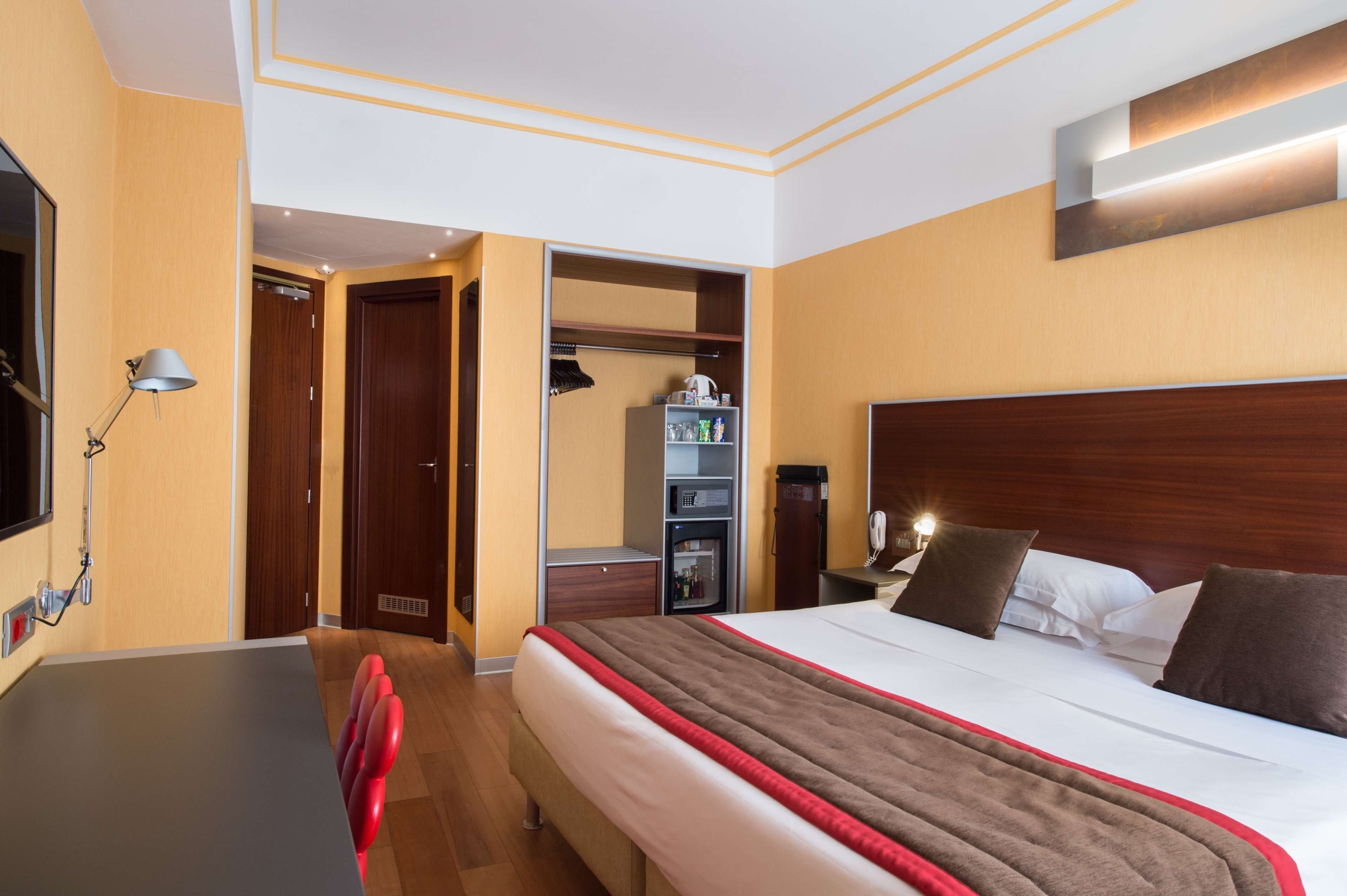A hotel room with a bed, bedside table, lamp, wardrobe, and a small desk with a chair.