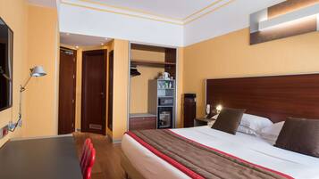 A hotel room with a bed, bedside table, lamp, wardrobe, and a small desk with a chair.