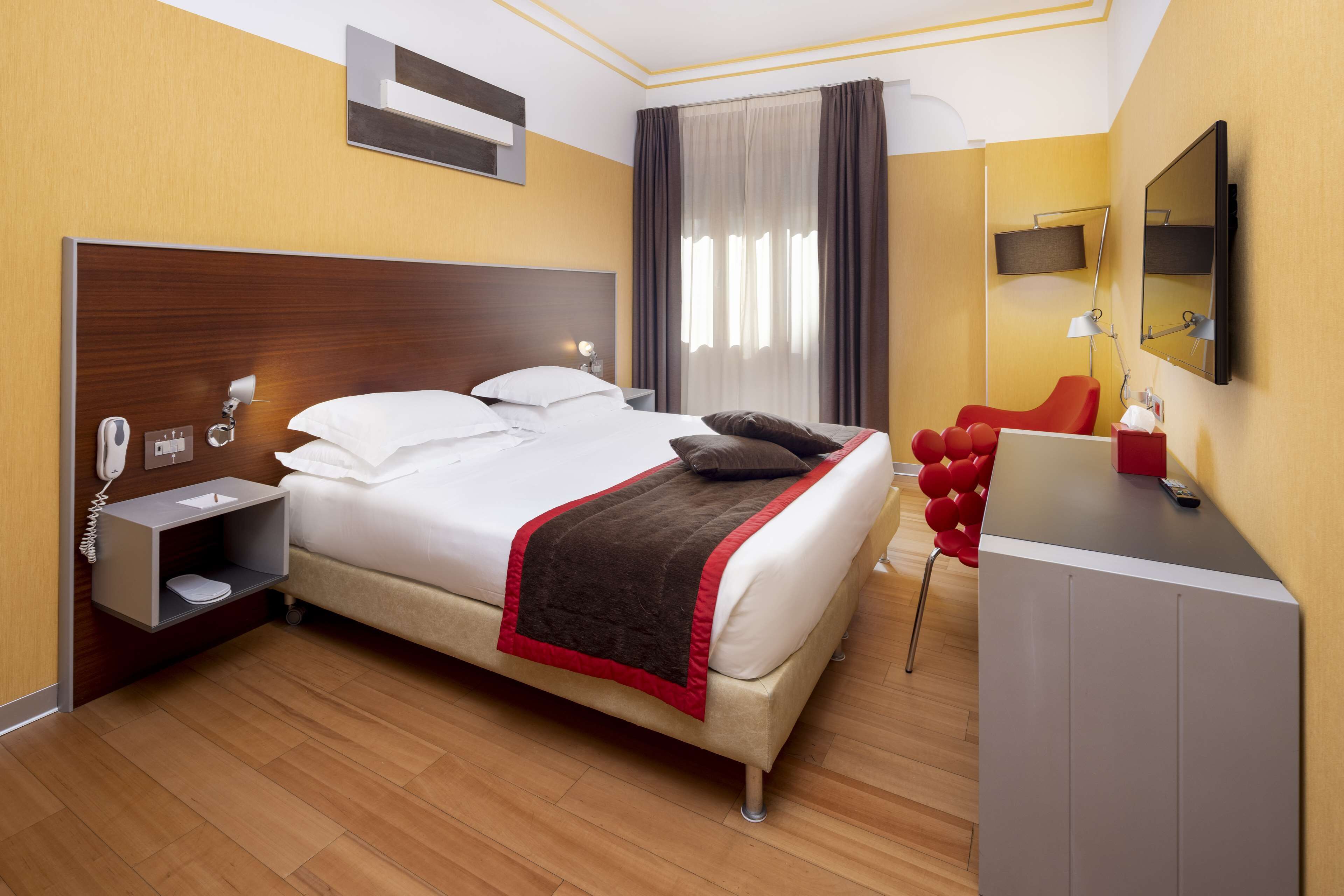 A hotel room with a large bed, bedside table with a lamp, a desk with a chair, and a TV mounted on the wall.