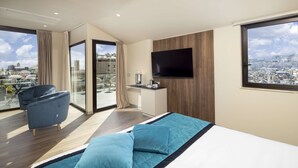 Habitación de hotel moderna con una cama grande, un televisor de pantalla plana, un balcón con vistas a la ciudad y una zona de estar con una silla.