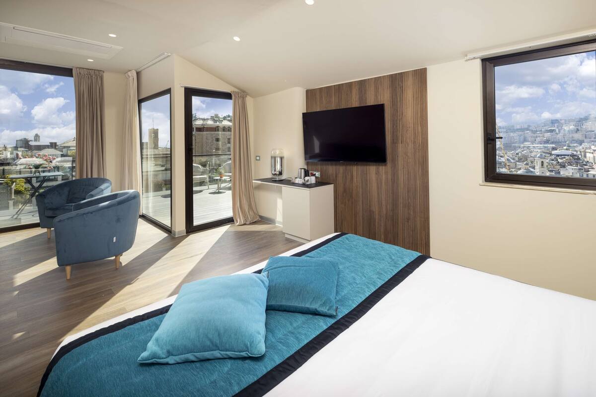 A modern hotel room with a large bed, a flat-screen TV, a balcony with a city view, and a seating area with a chair.