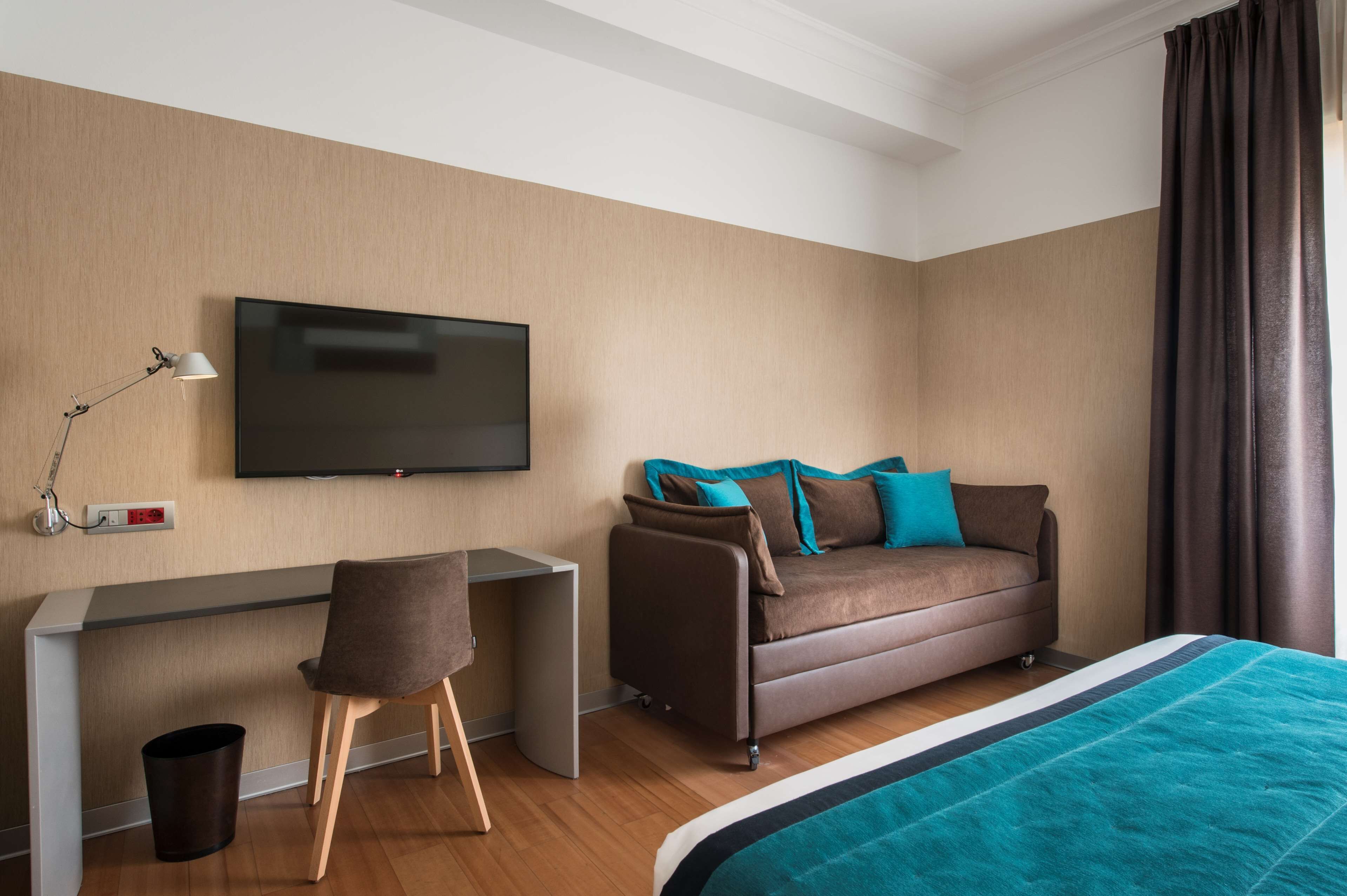Ein modernes Hotelzimmer mit einem Wand-Fernseher, einem Sofa mit petrolfarbenen und braunen Kissen, einem Schreibtisch mit Stuhl und einem Bett mit petrolfarbener Decke.