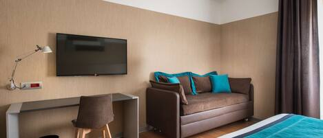 Una habitación de hotel moderna con un televisor de pantalla plana montado en la pared, un sofá con cojines azul verdosos y marrones, un escritorio con una silla y una cama con una manta azul verdosa.