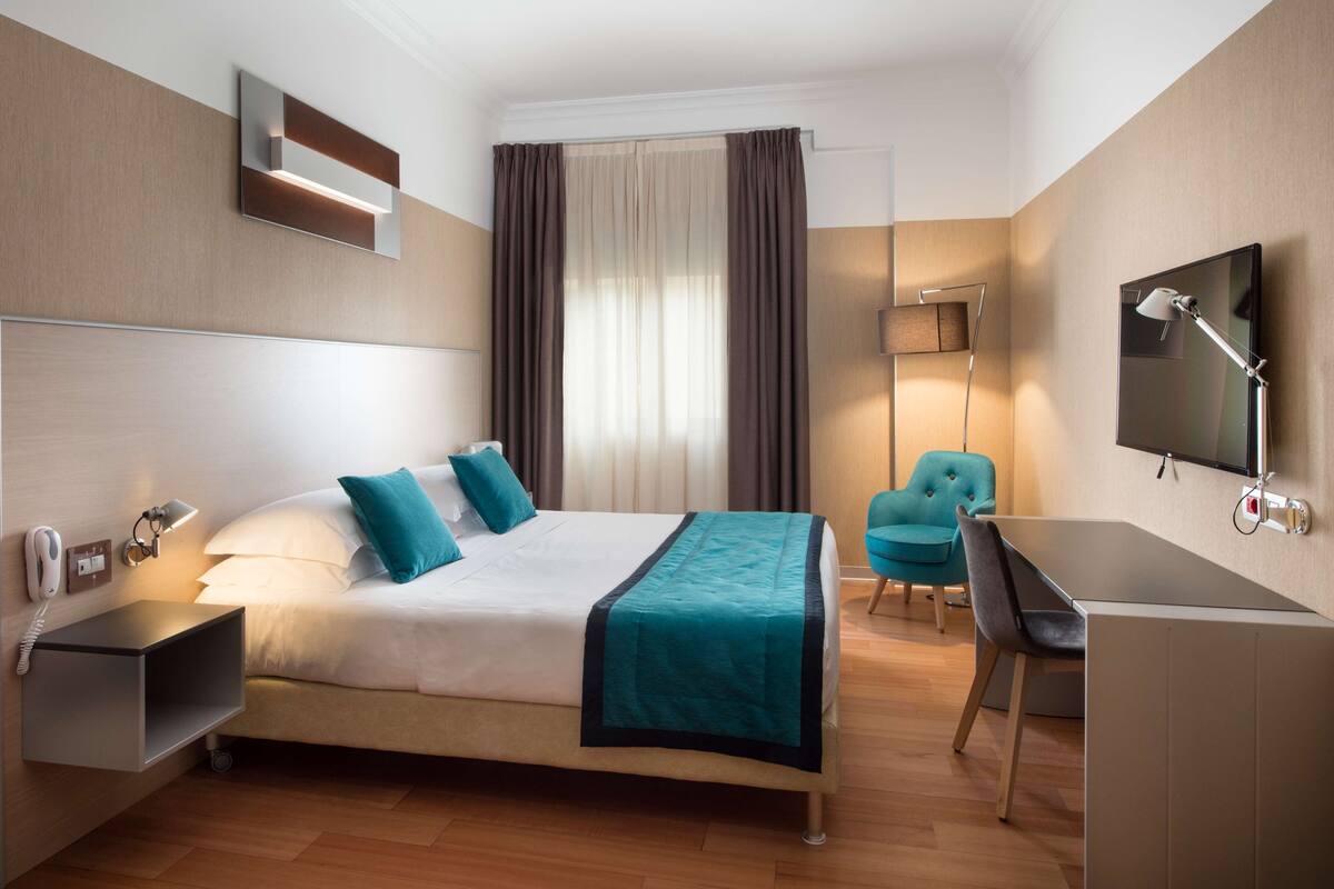A hotel room with a bed, a desk, a chair, a TV, and a bedside table with a lamp.
