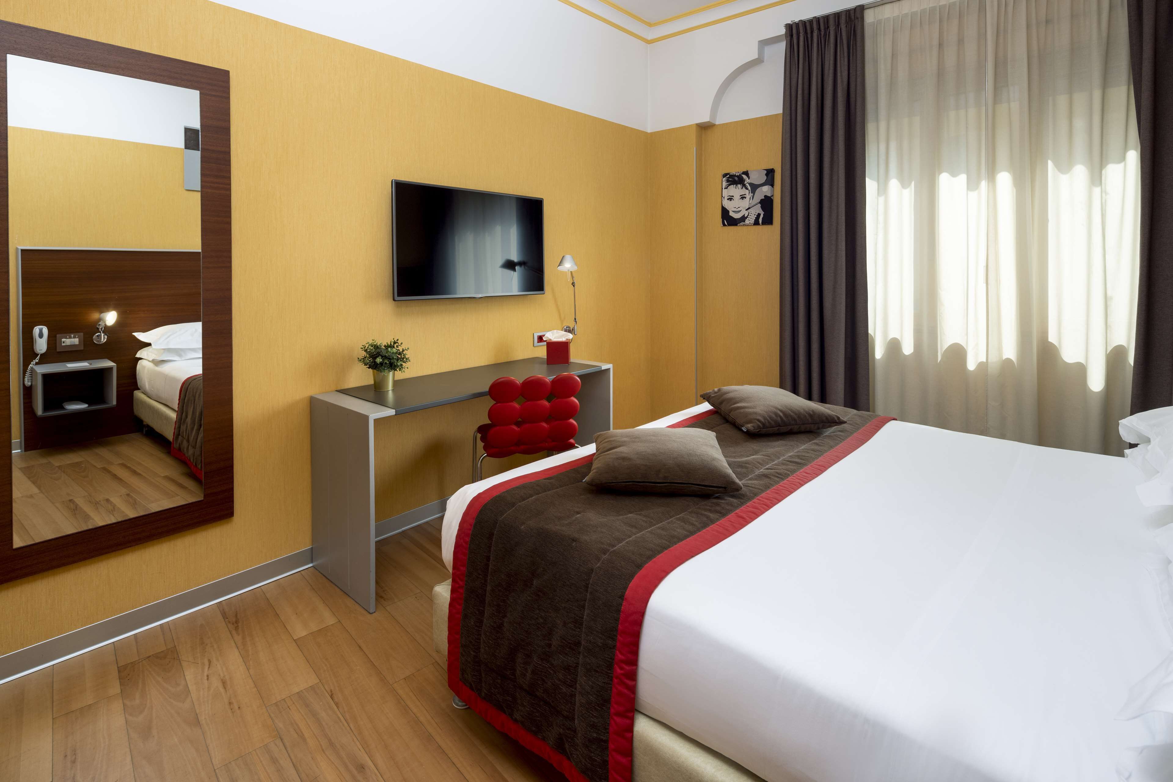 A hotel room with a bed, a desk, a TV, a mirror, and a window with curtains.