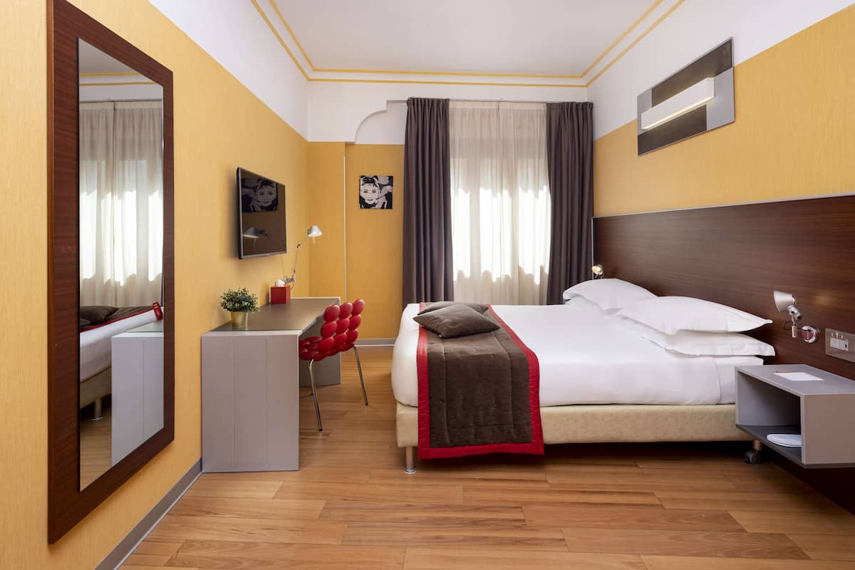A modern hotel room with a bed, bedside table, desk, chair, and a large mirror.