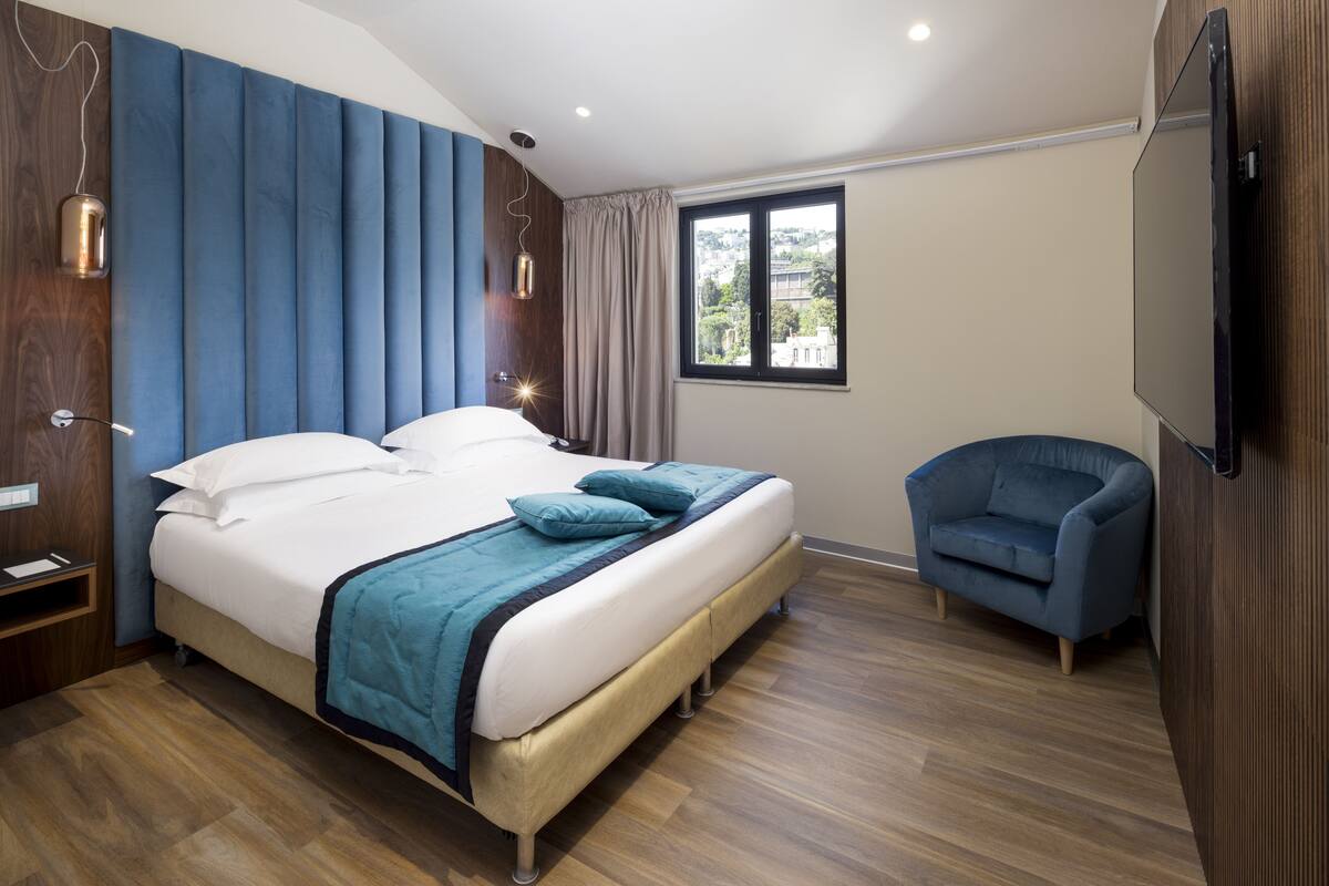 A modern hotel room with a large bed, a blue armchair, and a window with a city view.