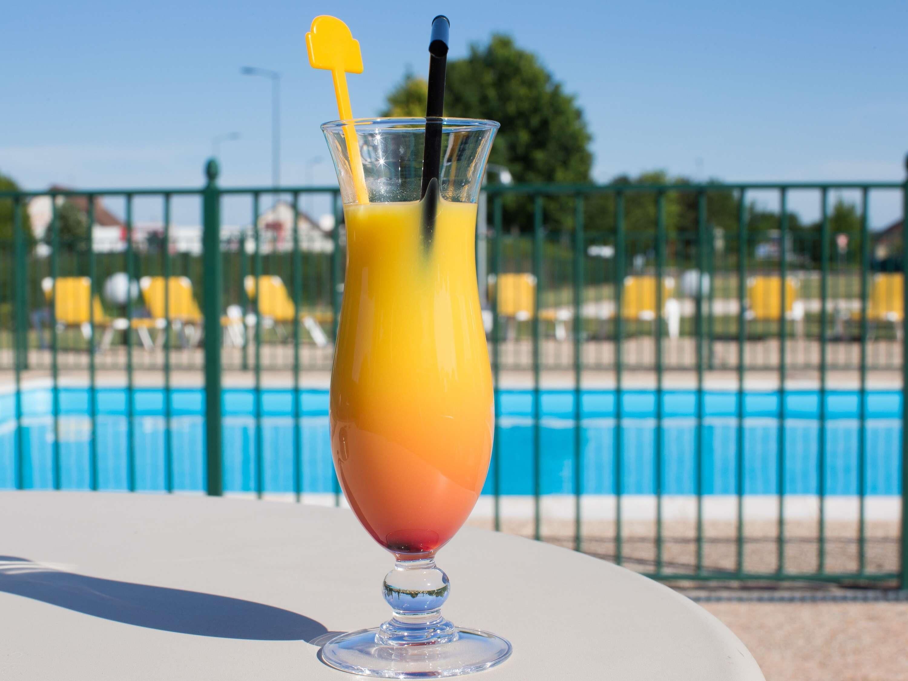 A glass of orange juice with a yellow and black straw on a table by a pool.