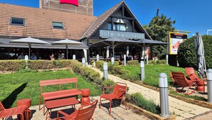 Un área de estar al aire libre con sillas y mesas rojas, un edificio con el letrero 'ibis' y un restaurante llamado 'Lefermeauxvins'.