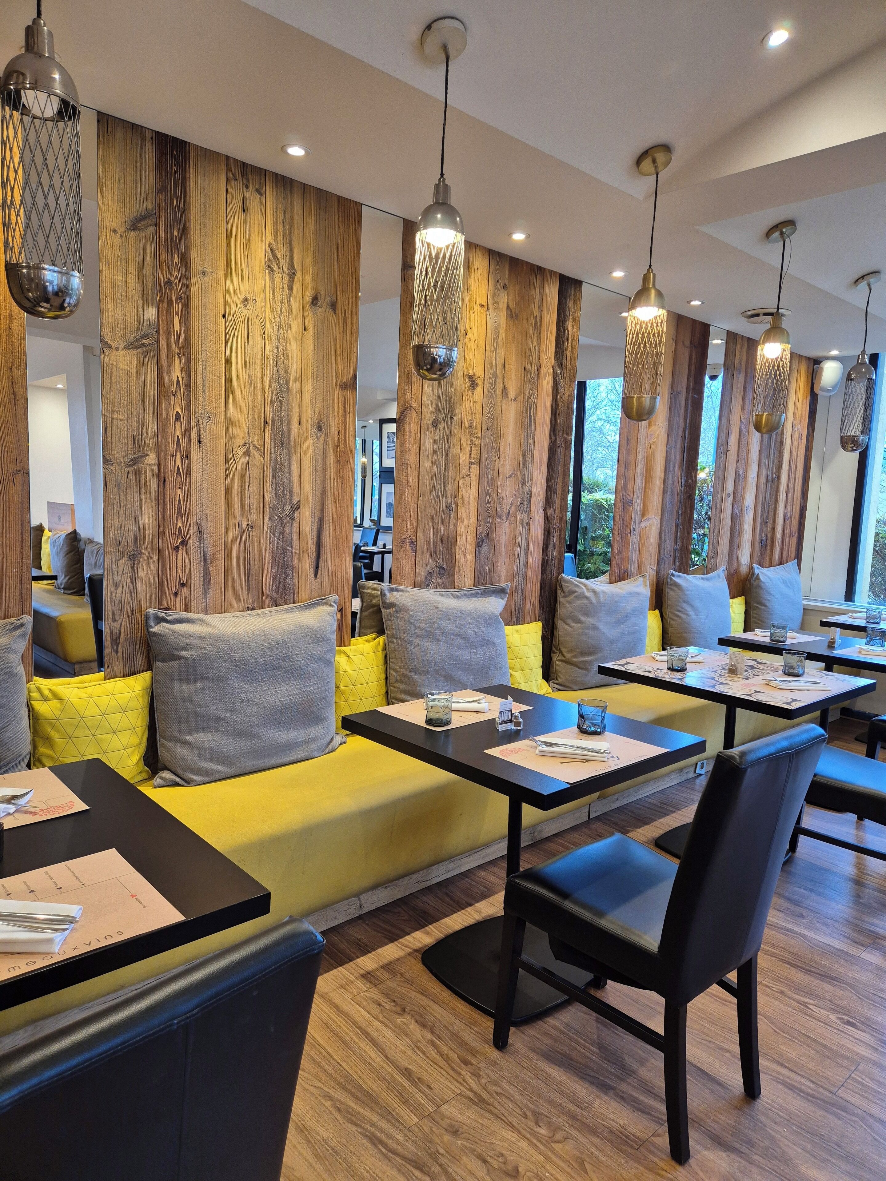 Ein modernes Restaurant mit Holzwandverkleidung, gelb gepolsterten Sitzgelegenheiten, schwarzen Tischen und hängenden Pendelleuchten.