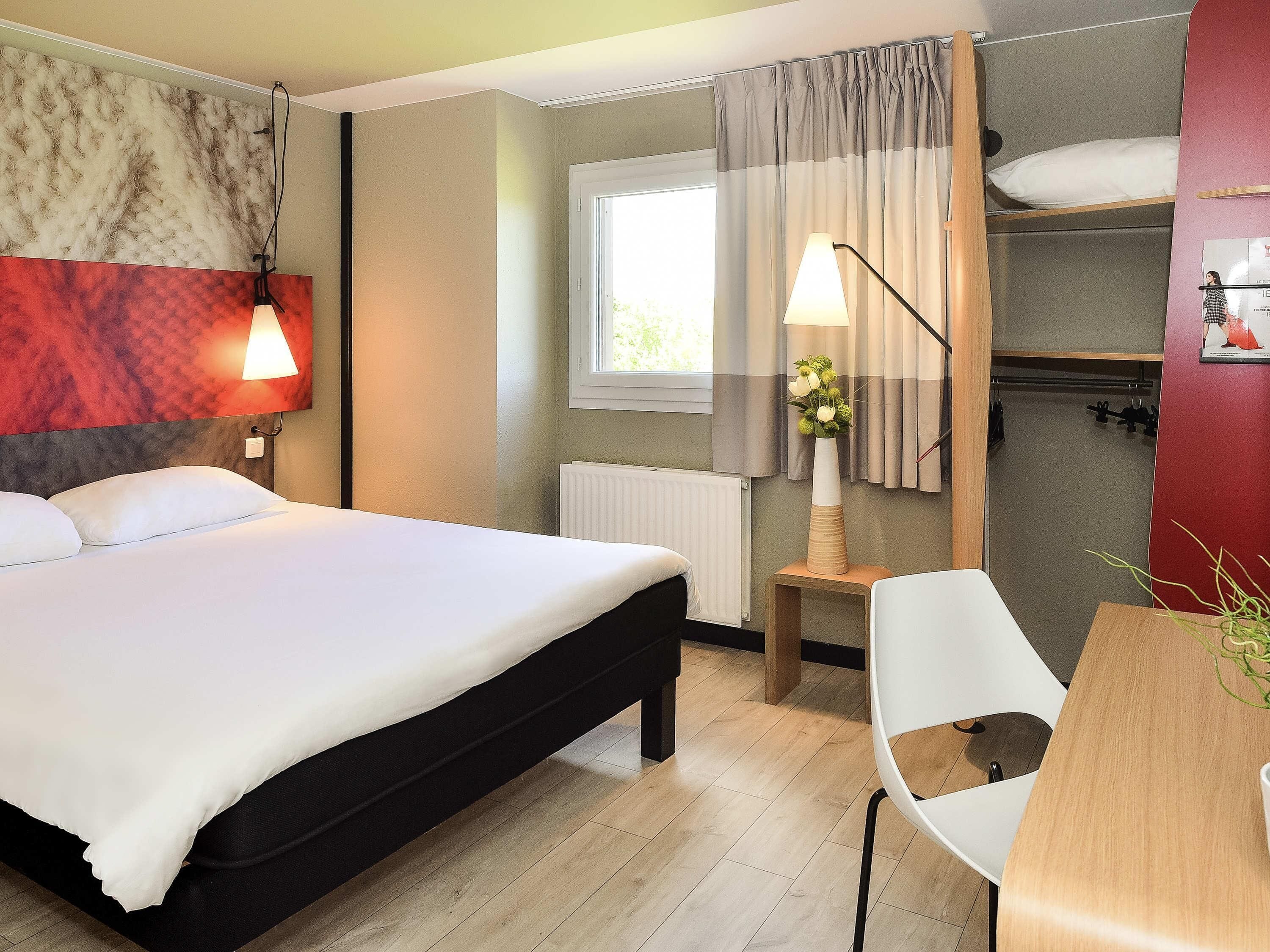 Habitación de hotel con cama, lámparas de noche, ventana con cortinas, radiador, mesa con silla y estante en la pared.