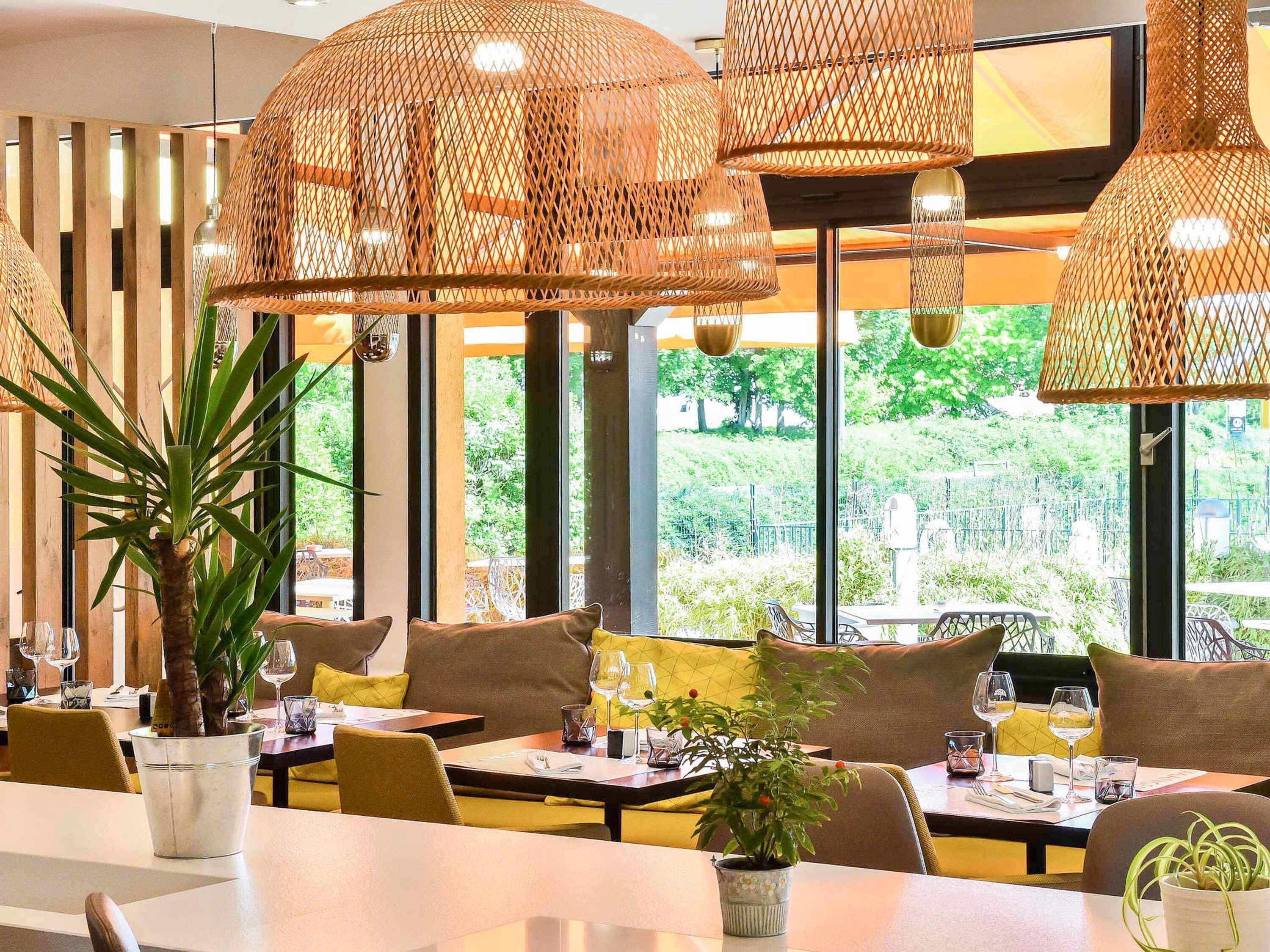 L’intérieur d’un restaurant, avec ses tables, ses chaises, ses lampes en osier suspendues et ses grandes fenêtres donnant sur un paysage verdoyant.