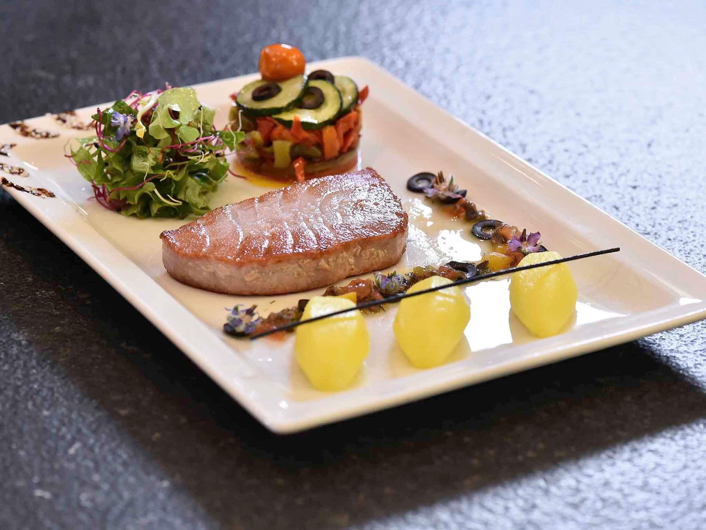 A square plate with a grilled meat, a vegetable terrine, a green salad, and three small yellow items.