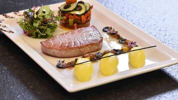 A square plate with a grilled meat, a vegetable terrine, a green salad, and three small yellow items.
