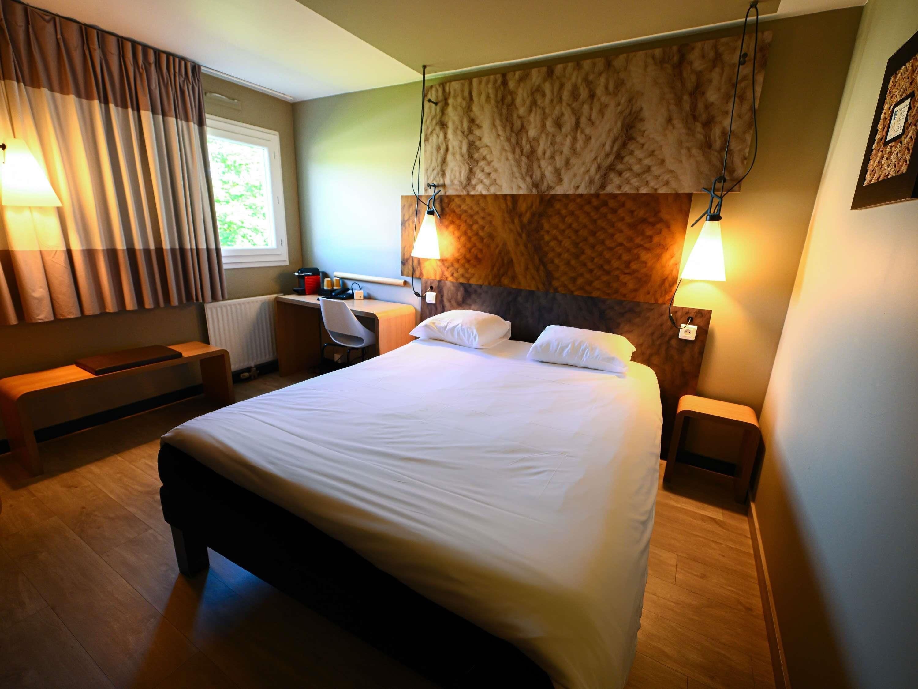 Quarto de hotel com uma cama grande, mesas de cabeceira, uma escrivaninha com cadeira, janela com cortinas e uma luminária na parede.