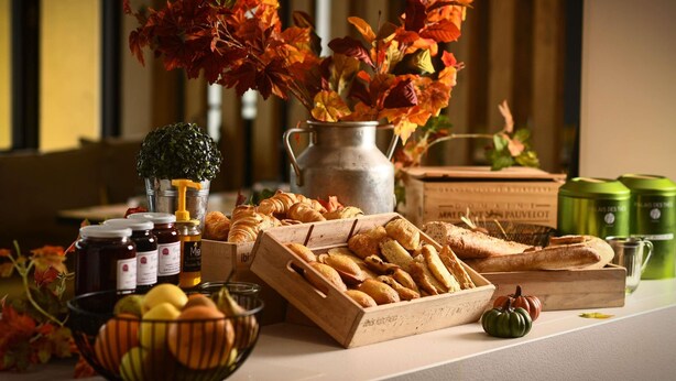 Una mesa con pasteles, un bol de fruta y un florero con hojas de otoño.