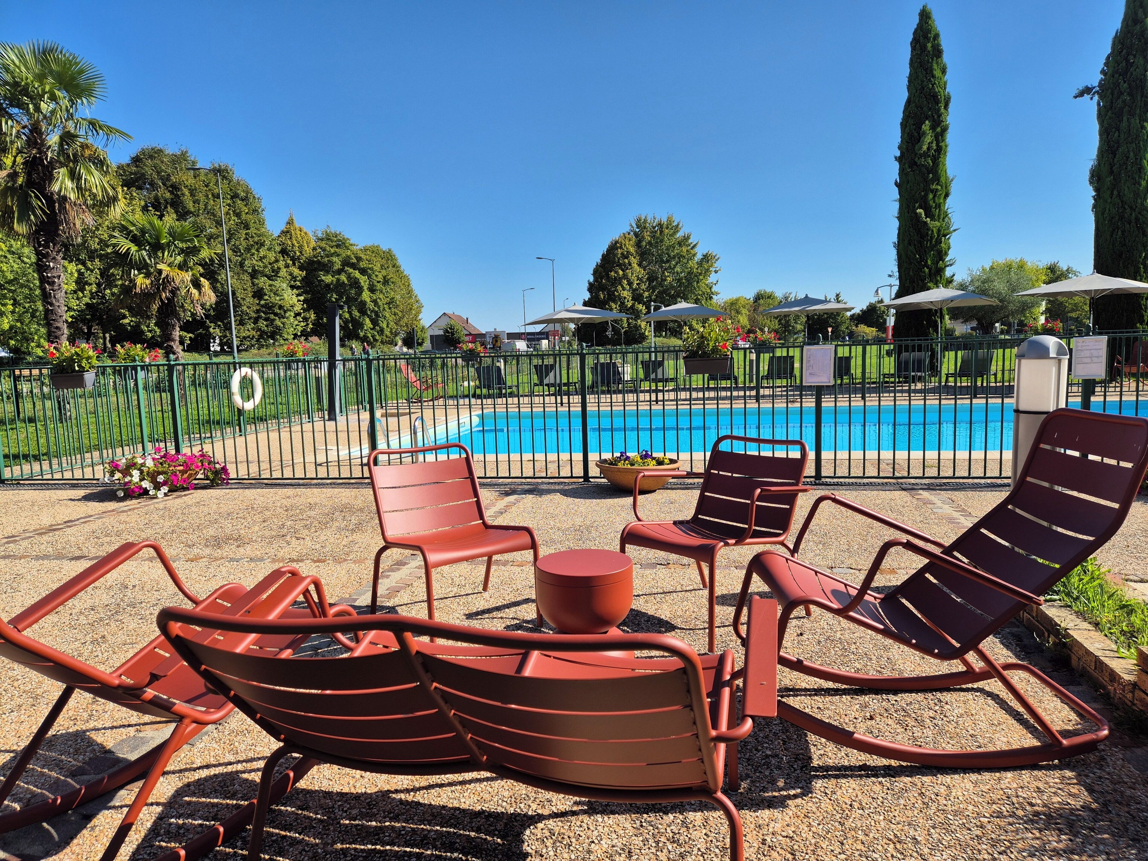 Zona junto a la piscina con sillas reclinables rojas y una mesita, rodeada por una valla metálica y follaje.