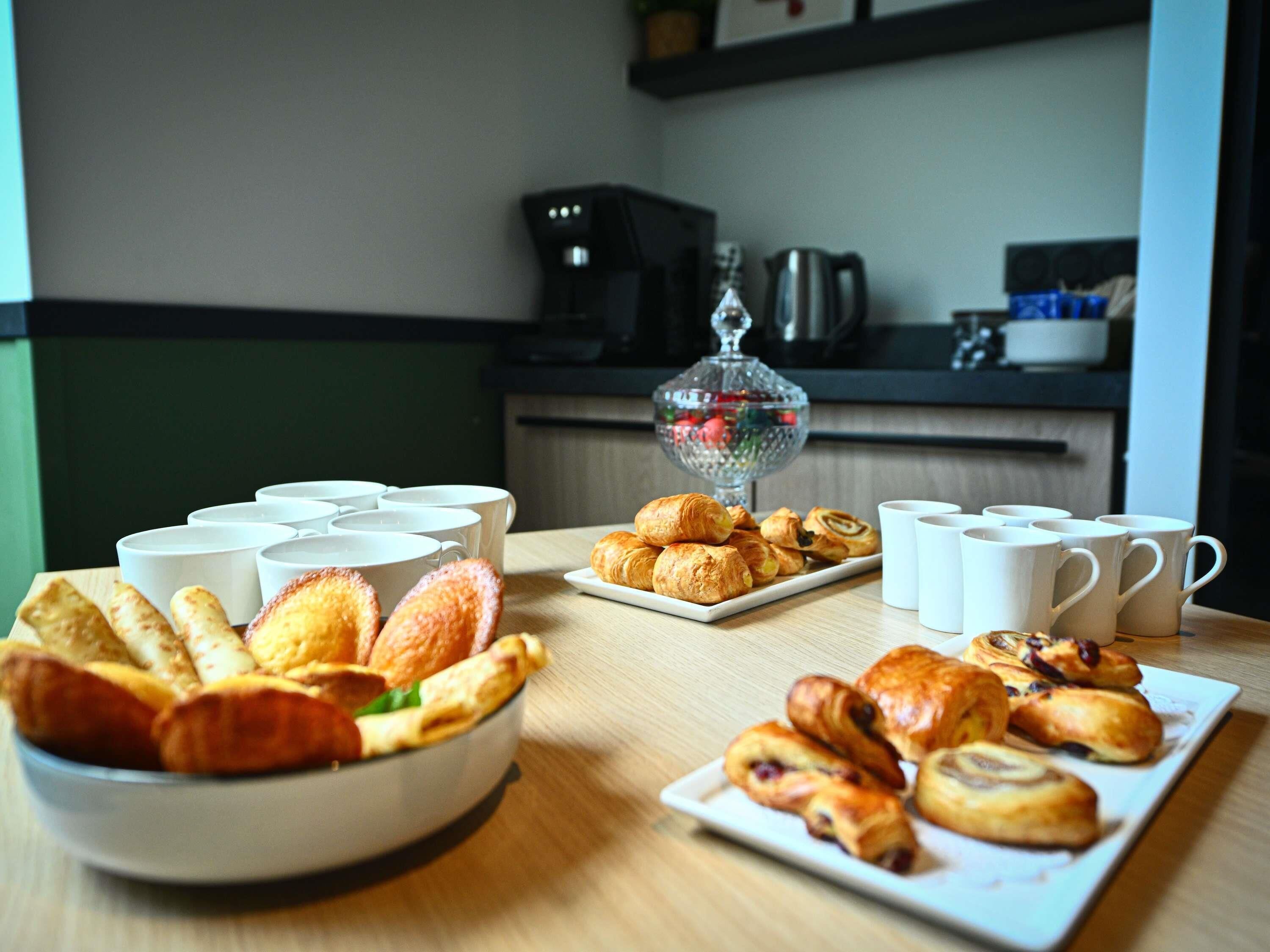 Een ontbijtbuffet met gebak, croissants en muffins, plus verschillende witte bekers en een koffiezetapparaat op de achtergrond.