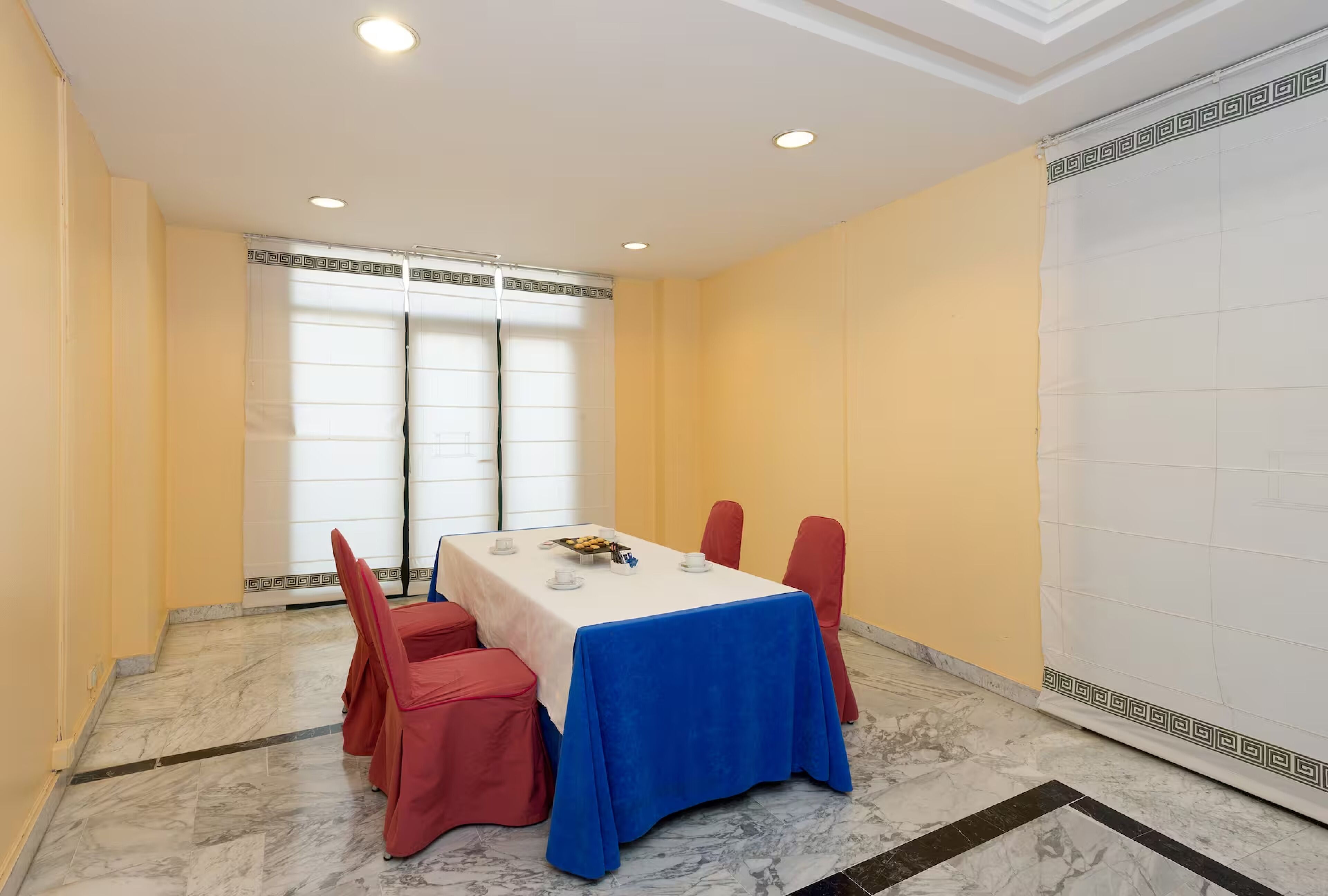 A conference room with a marble floor, a white table with a blue tablecloth, red chairs, and a large window with blinds.