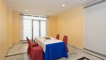Un salón de conferencias con piso de mármol, una mesa blanca cubierta con mantel azul, sillas rojas y un ventanal con persianas.