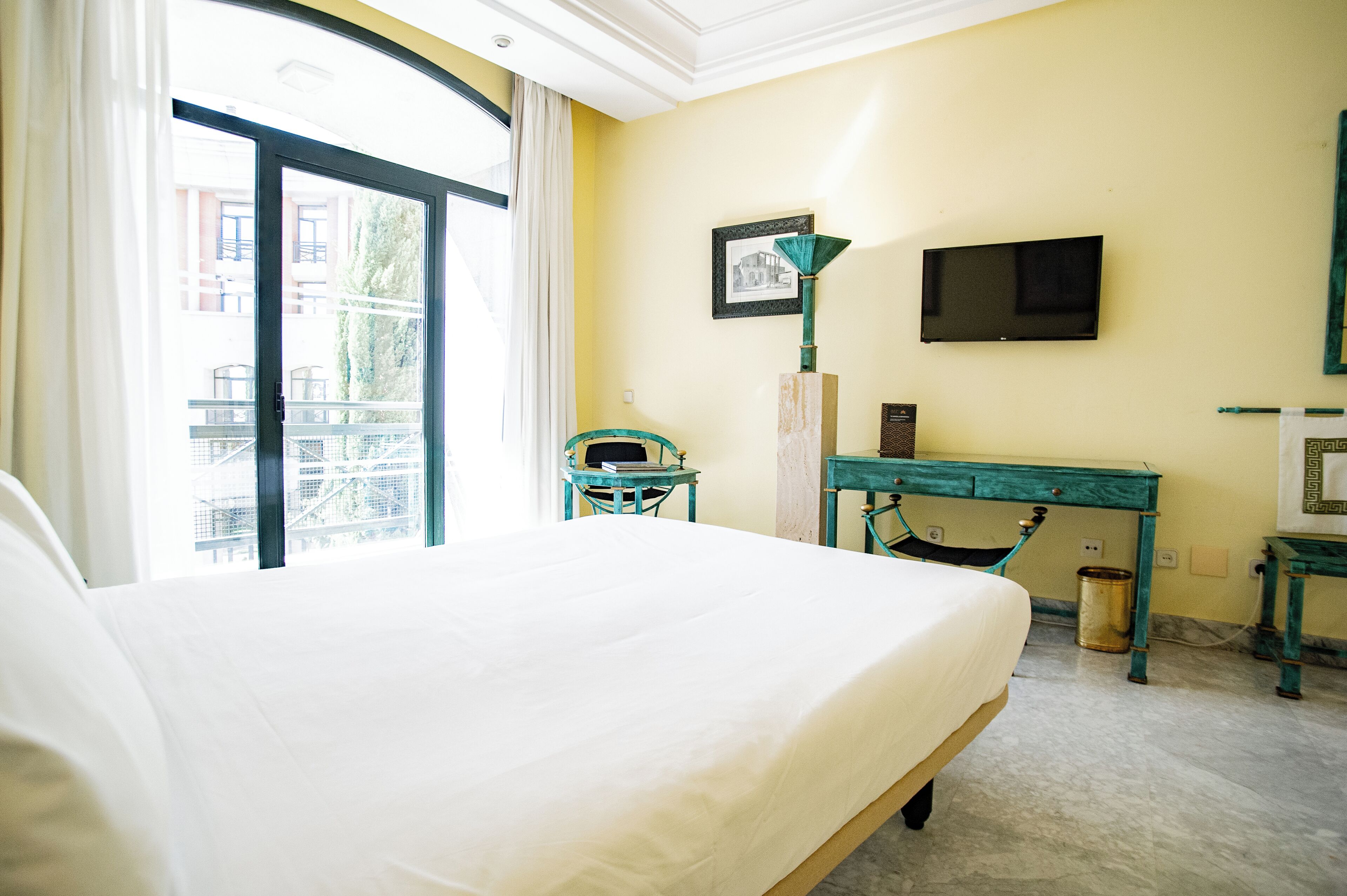 A hotel room with a bed, a desk, a television, and a window with a view of buildings.