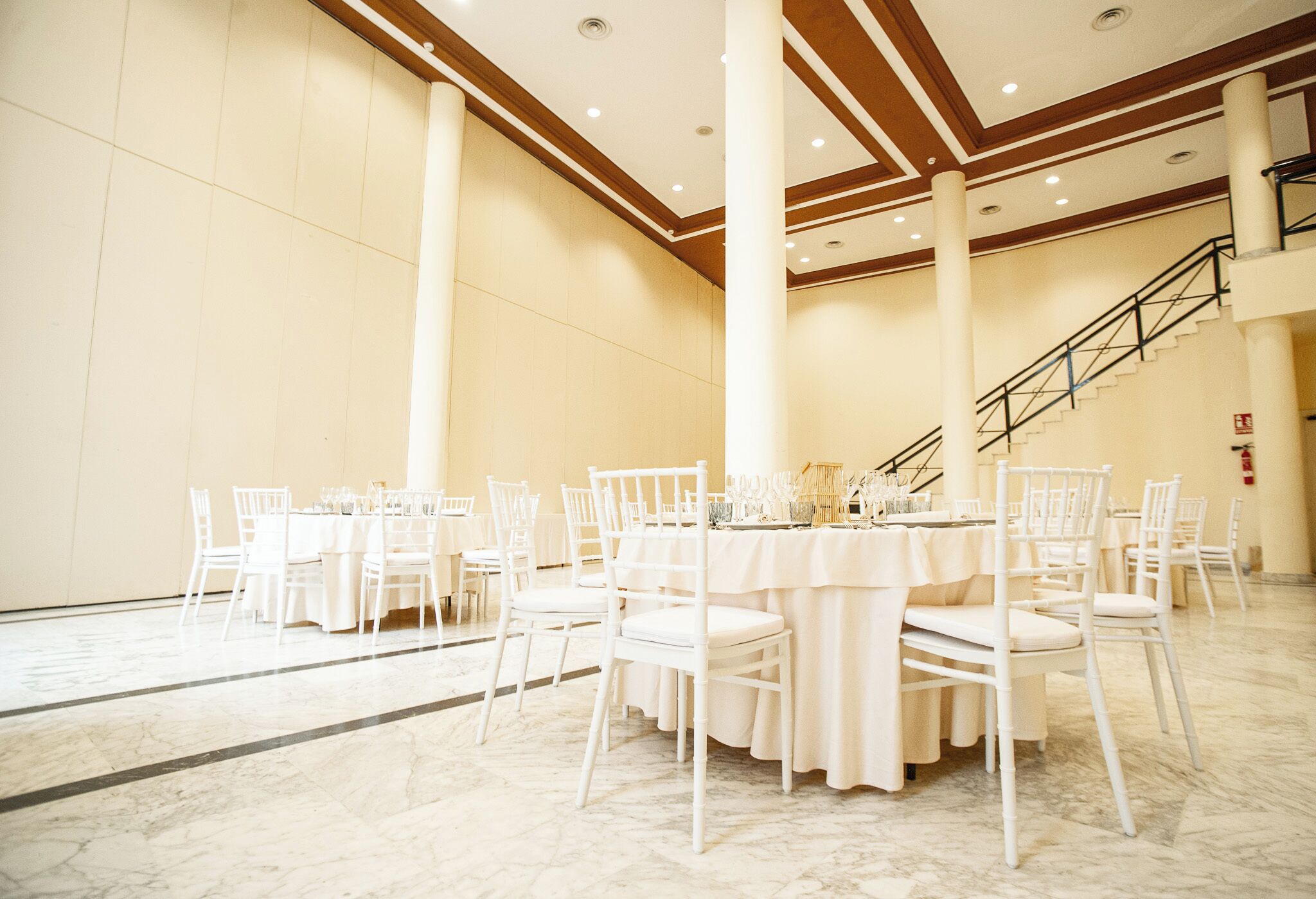 A spacious hall with marble flooring, white tables and chairs, and a staircase in the background.