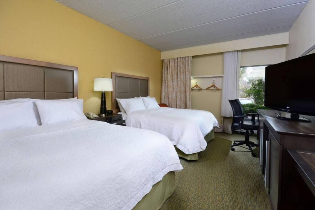 A hotel room with two beds, a desk with a computer, a chair, a TV, and a window with curtains.