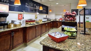 A kitchen area with wooden cabinets, granite countertops, and various kitchen appliances. There are hanging orange pendant lights, a coffee machine, and a display of fruits.