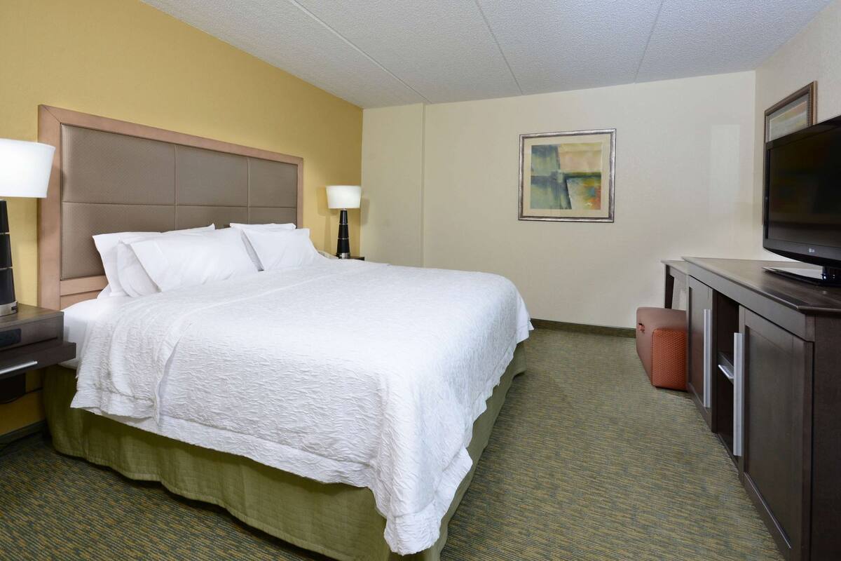 A hotel room with a bed, a television, a desk with a lamp, and a painting on the wall.