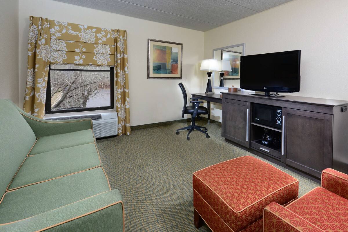 A hotel room with a green sofa, a red patterned ottoman, a desk with a lamp, a television, and a window with curtains.