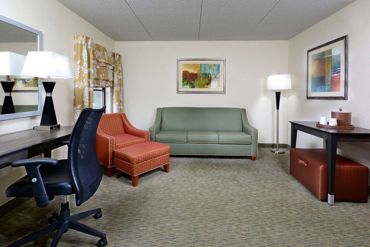 A hotel room with a desk, two chairs, a sofa, a lamp, a painting, and a suitcase.