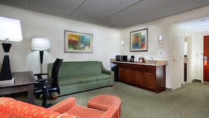 A hotel room with a green sofa, a red patterned chair, a desk with a lamp, a kitchenette, and artwork on the walls.