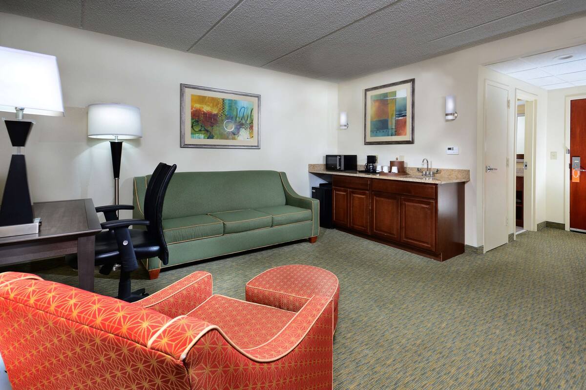 A hotel room with a green sofa, a red patterned chair, a desk with a lamp, a kitchenette, and artwork on the walls.