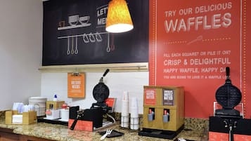 A kitchen area with waffle-making equipment, a waffle batter dispenser, and a sign promoting waffles.