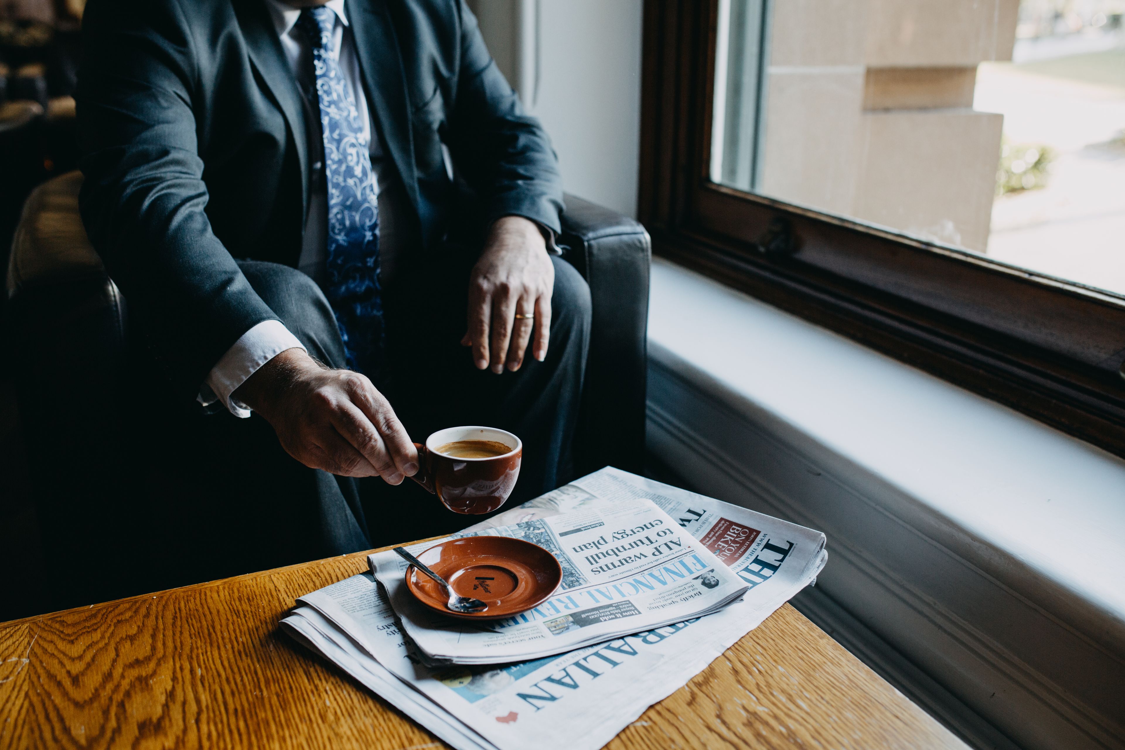 A person sitting by a window with a newspaper, a cup, and a spoon on a wooden table.