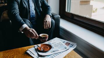 A person sitting by a window with a newspaper, a cup, and a spoon on a wooden table.