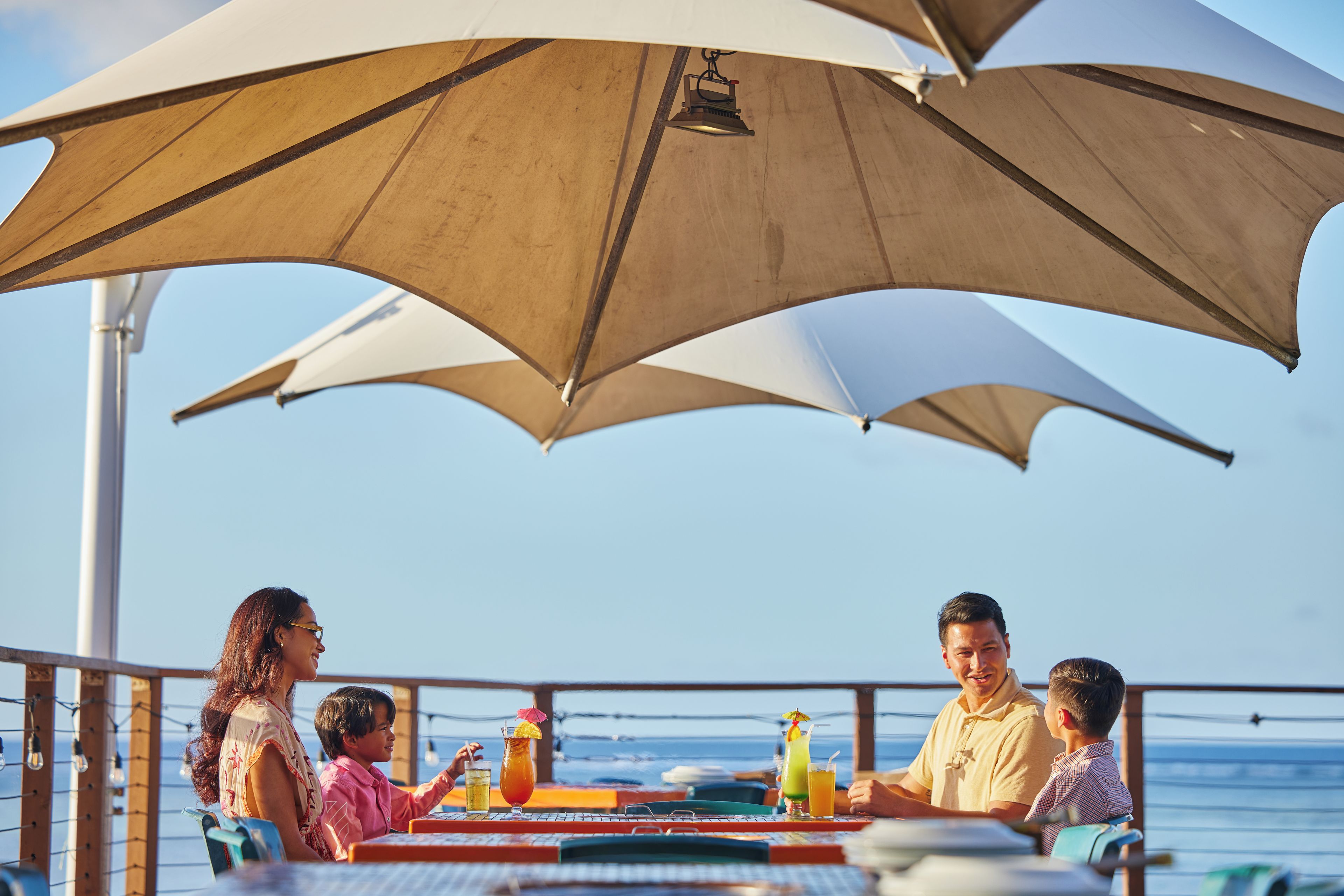 A family dining at an outdoor table with a view of the sea, under a large umbrella.