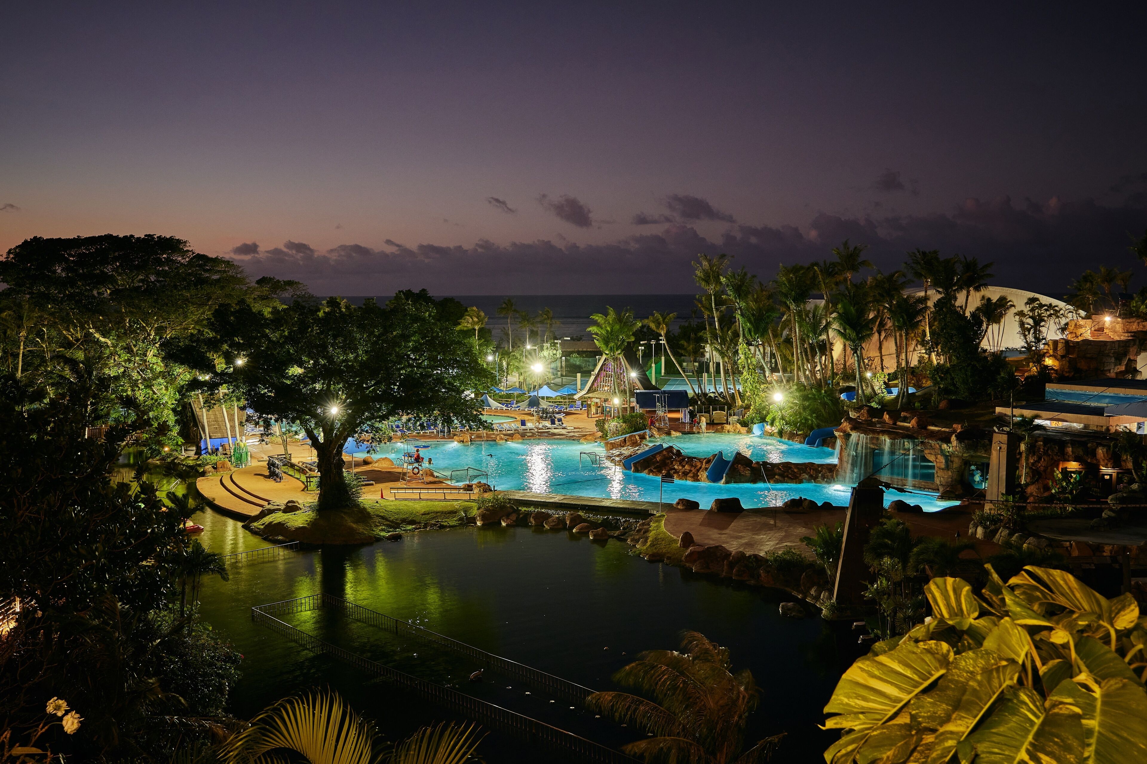 A nighttime view of a resort with a large swimming pool, palm trees, and a waterfall.
