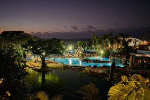 A nighttime view of a resort with a large swimming pool, palm trees, and a waterfall.