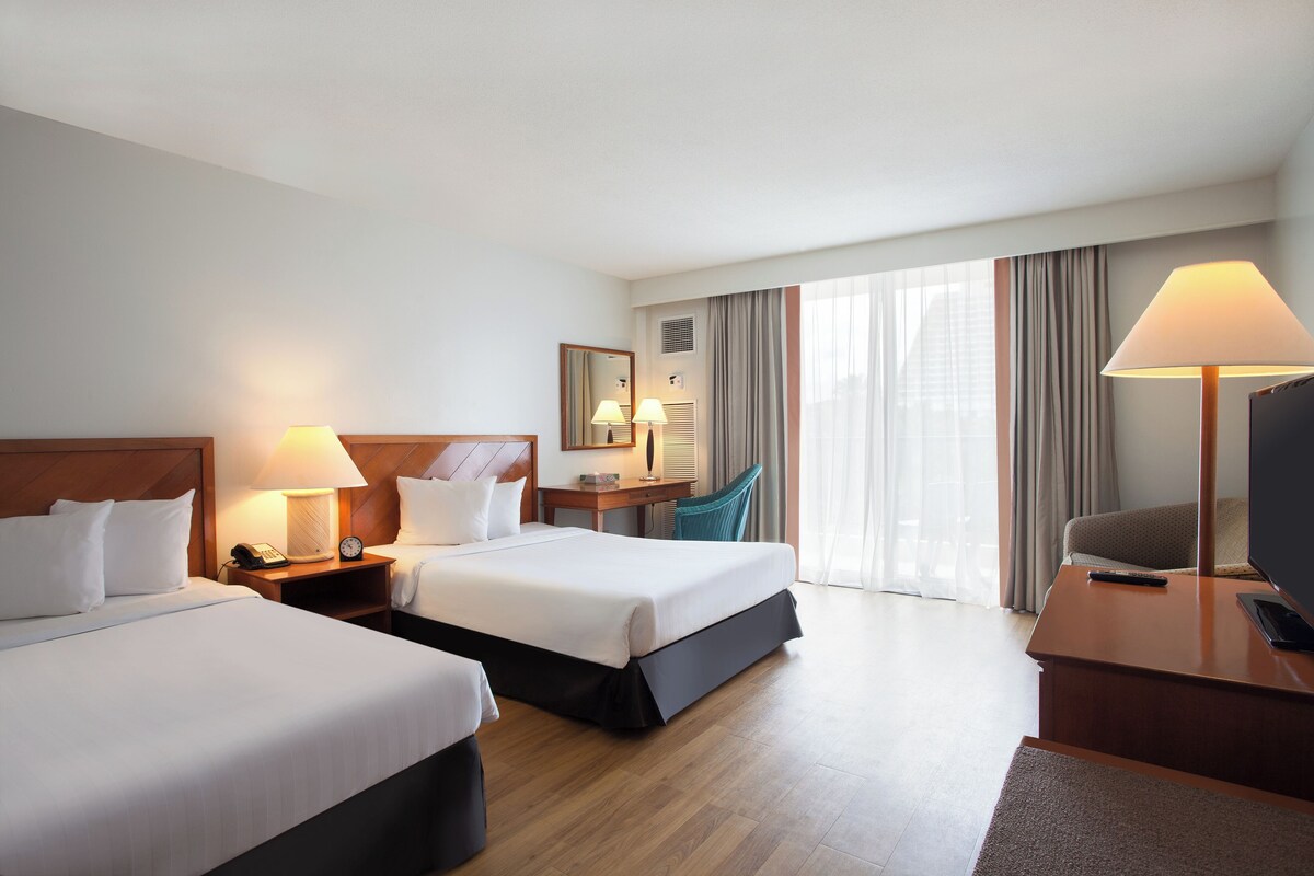 Habitación de hotel con dos camas, un escritorio, una silla, un televisor y un ventanal con cortinas.