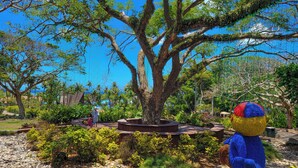 A large tree with a circular stone bench in the center, surrounded by various plants and a decorative sculpture.