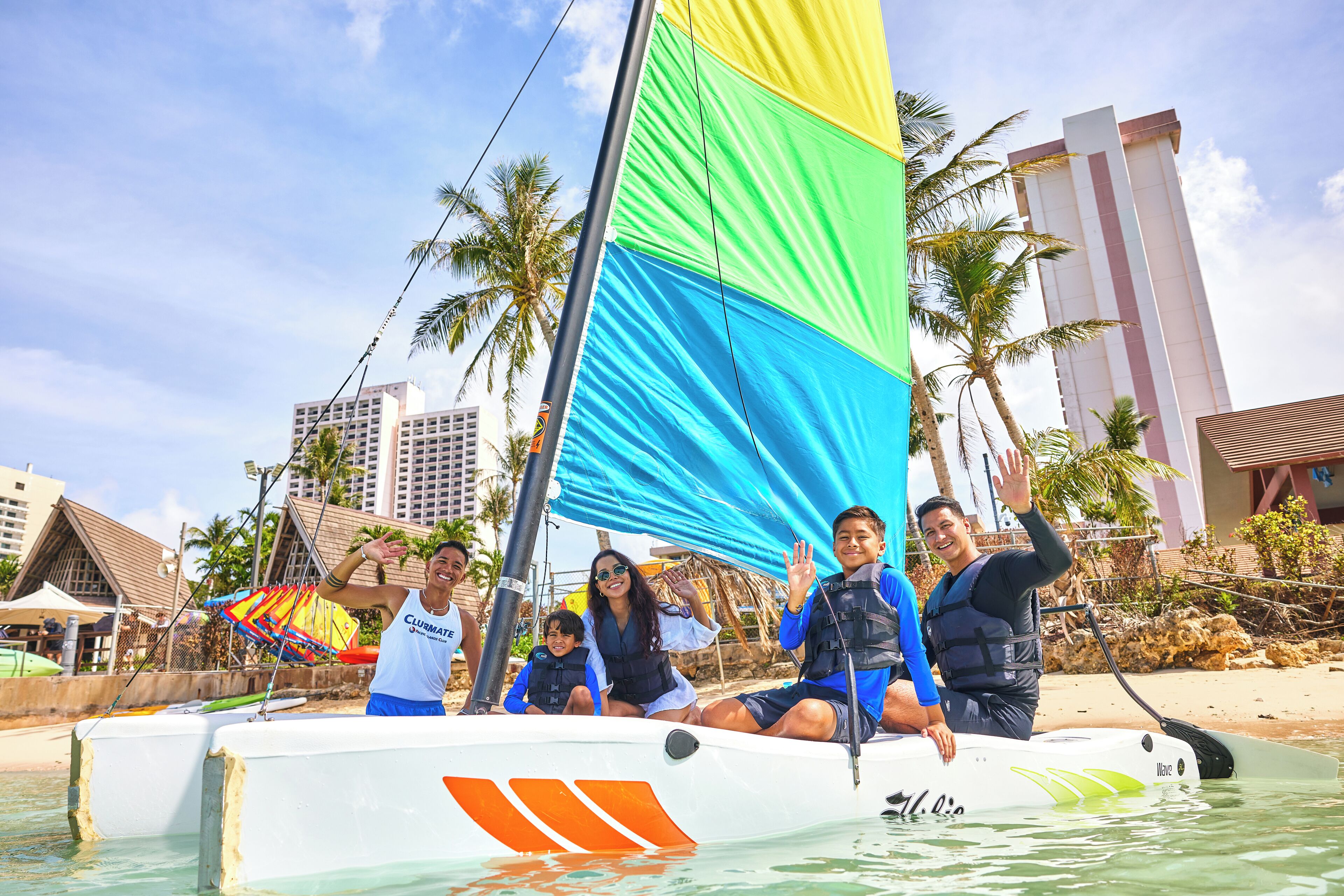 A sailboat with a colorful sail, people on board, and a beach with palm trees and buildings in the background.